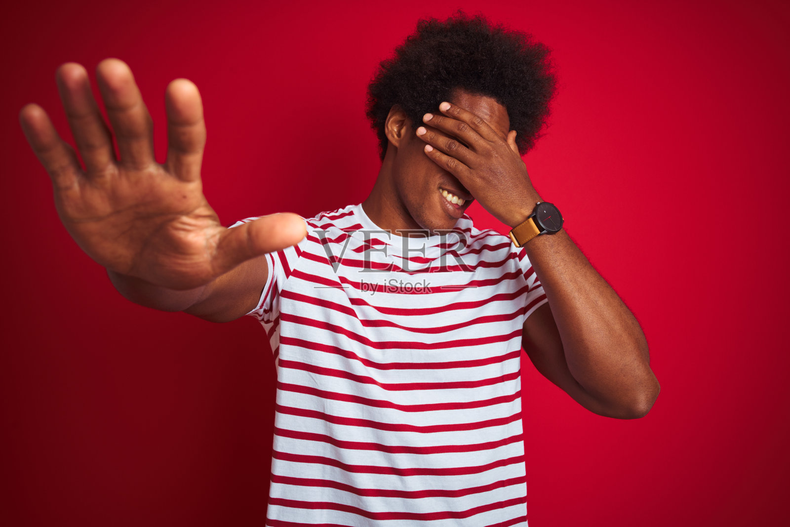 年轻的非洲裔美国人与非洲式头发穿条纹t恤在孤立的红色背景覆盖眼睛的手和做停止手势与悲伤和恐惧的表情。尴尬和消极的概念。照片摄影图片