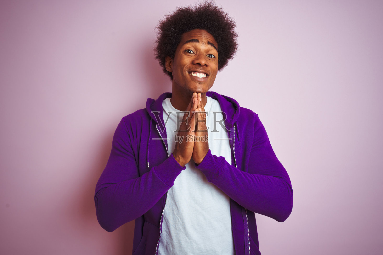 年轻的非洲裔美国人穿着紫色的运动衫站在孤立的粉红色背景与手一起祈祷，请求原谅自信的微笑。照片摄影图片