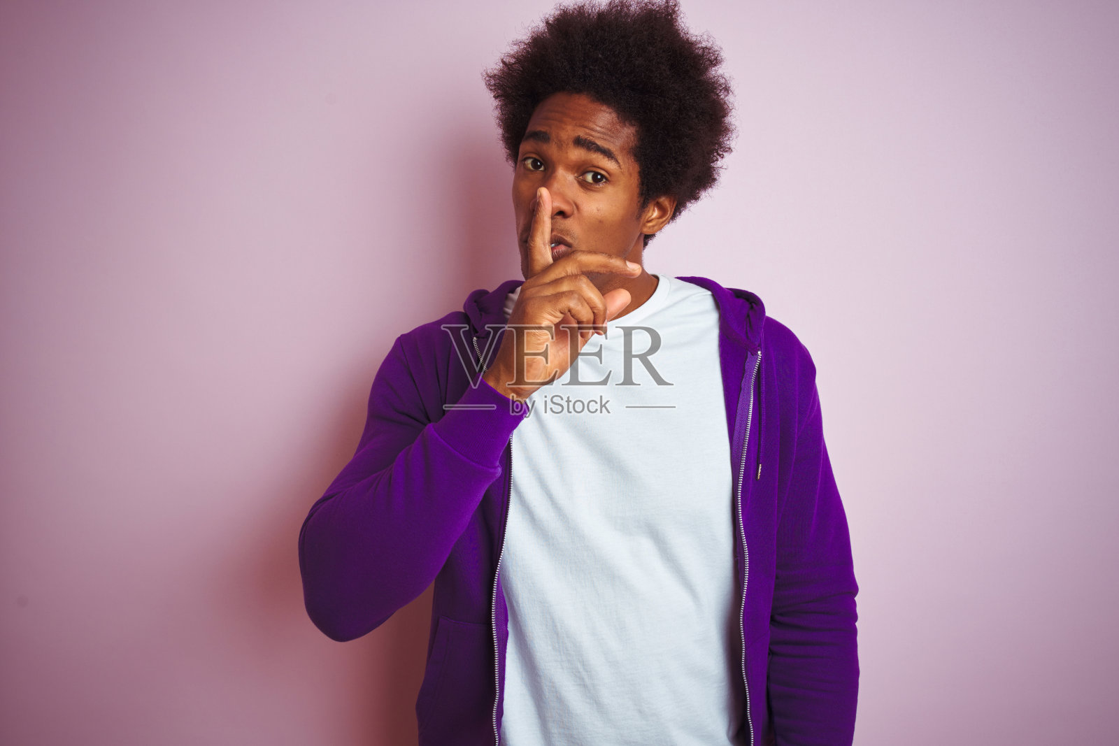 年轻的非洲裔美国人穿着紫色运动衫站在孤立的粉红色背景要求安静与手指在嘴唇上。沉默与秘密的概念。照片摄影图片