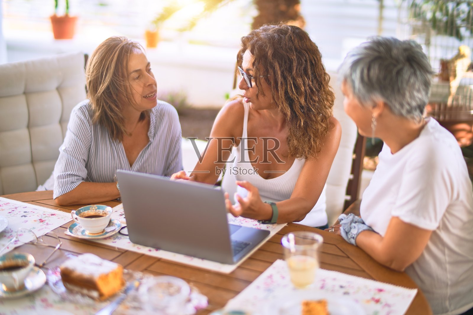 遇见中年妇女吃午饭喝咖啡。在一个阳光明媚的日子里，成熟的朋友们在家里用笔记本电脑，微笑着照片摄影图片