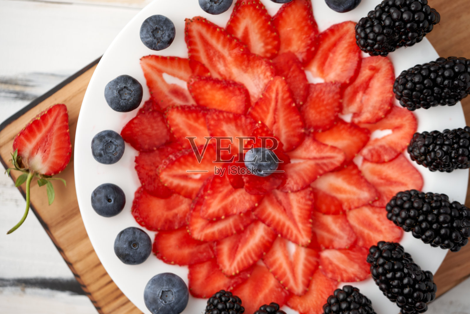 切成两半的有机草莓、蓝莓和黑莓，对称地摆放在砧板上的一个白色盘子上，背景是浅色的木头。饮食，素食菜单。从上面的观点照片摄影图片
