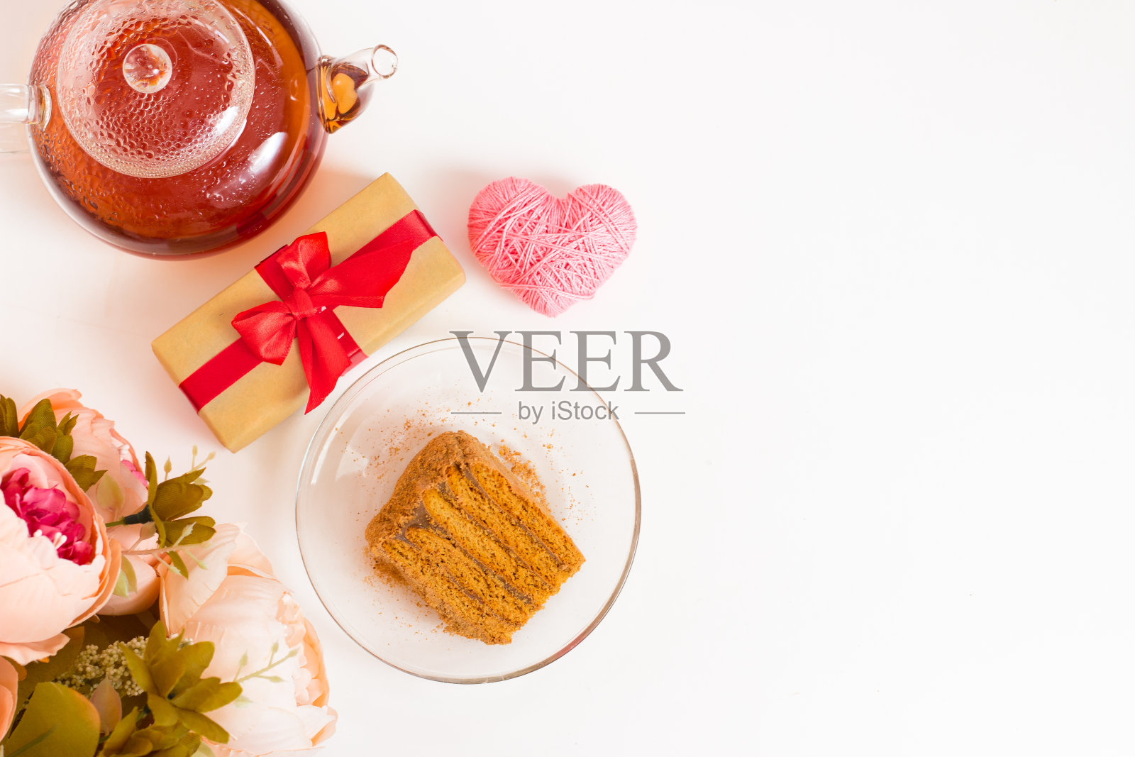 白色桌子上的玻璃杯里装着红茶。透明碟上的一片蜂蜜蛋糕。一束粉红色的花和一个带红色蝴蝶结的礼物。国际妇女节。情人节。照片摄影图片