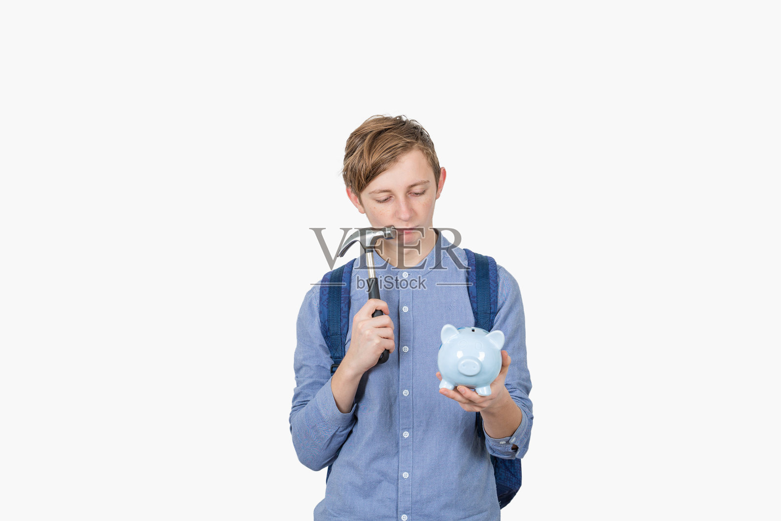 沮丧的学生男孩有怀疑打破他的小猪银行使用一个锤子孤立在白色的背景。存款少，没有足够的钱来签学习合同。照片摄影图片