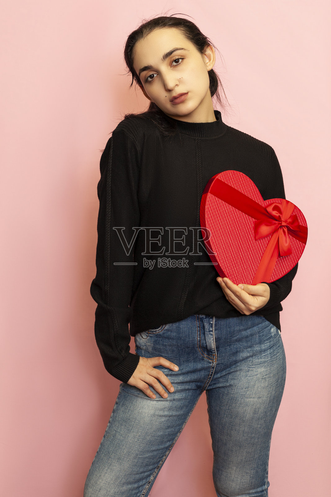 手捧心形礼盒的中东年轻女性：穿着休闲服装，手拿红色礼品盒的迷人时尚模特。情人节礼物，周年纪念日，生日照片摄影图片