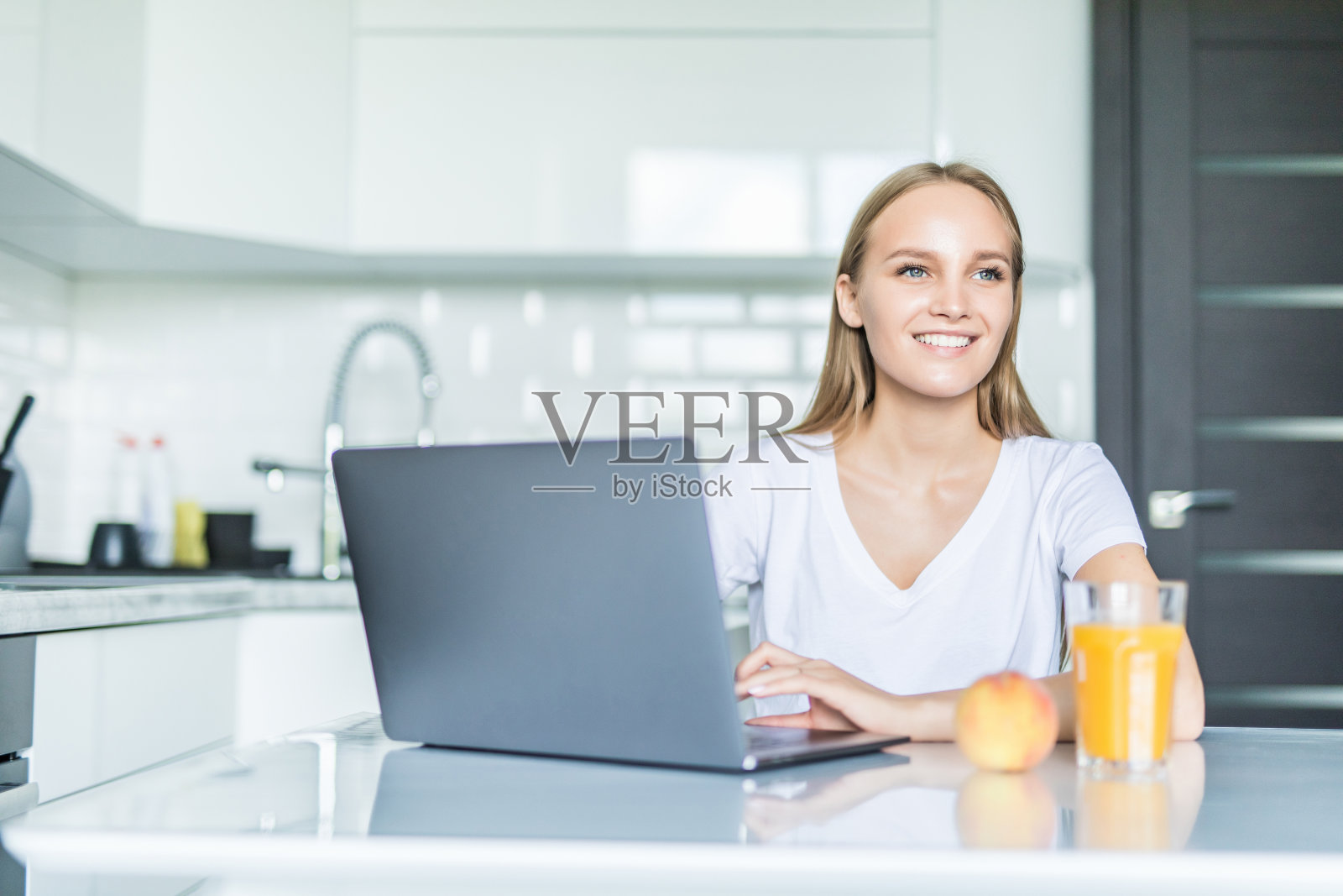 在厨房里一边用笔记本电脑一边喝橙汁的美丽女人照片摄影图片