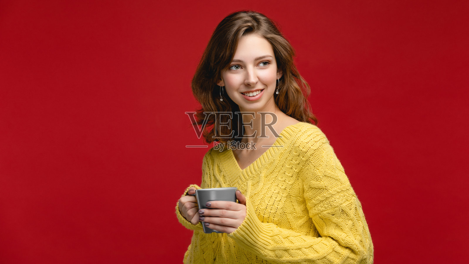 红色背景的演播室里穿着黄色套头衫的年轻女子。概念照片与一杯饮料照片摄影图片