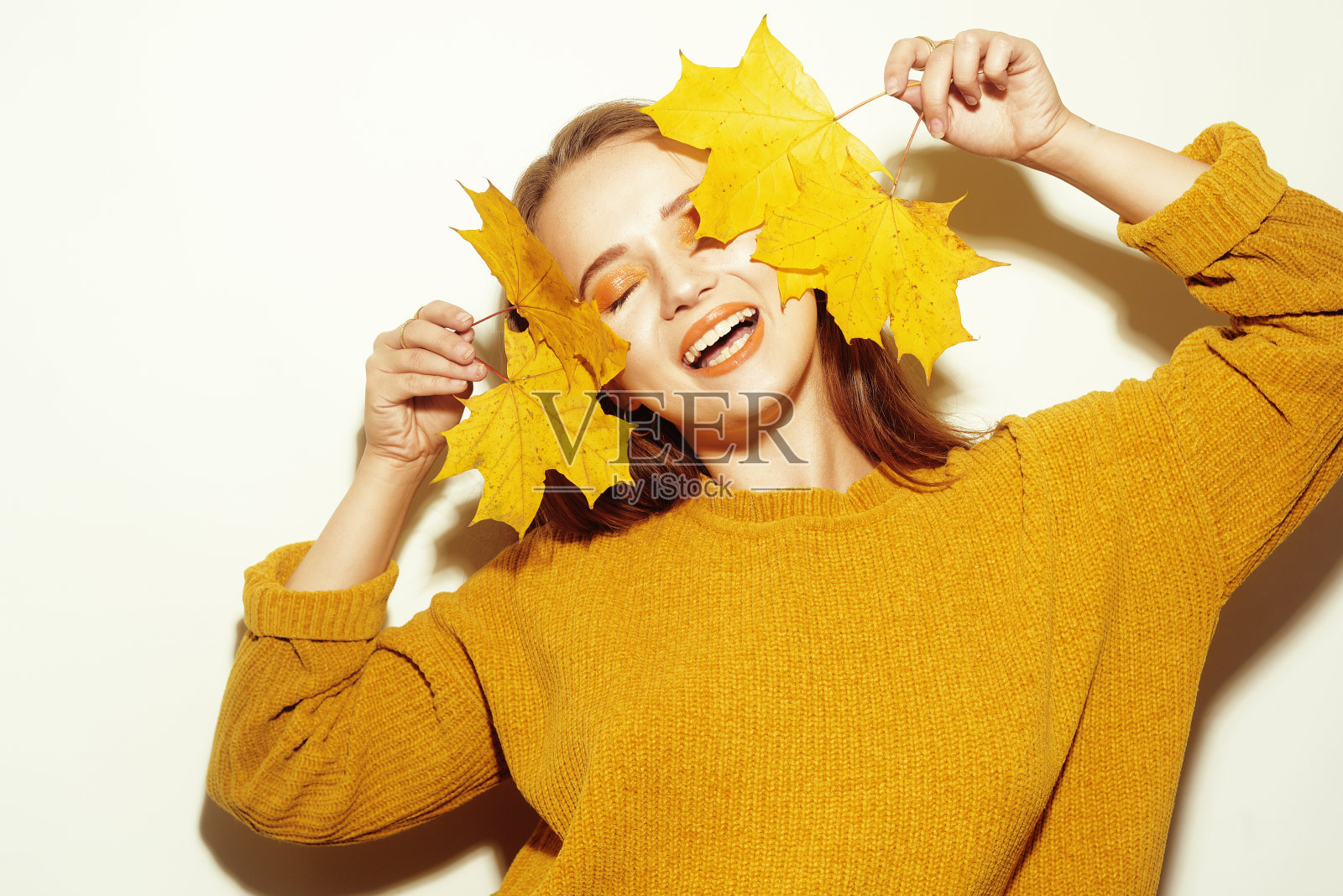 身穿黄色秋季毛衣的女孩在树叶中玩耍，笑容满面，用树叶遮住脸庞照片摄影图片