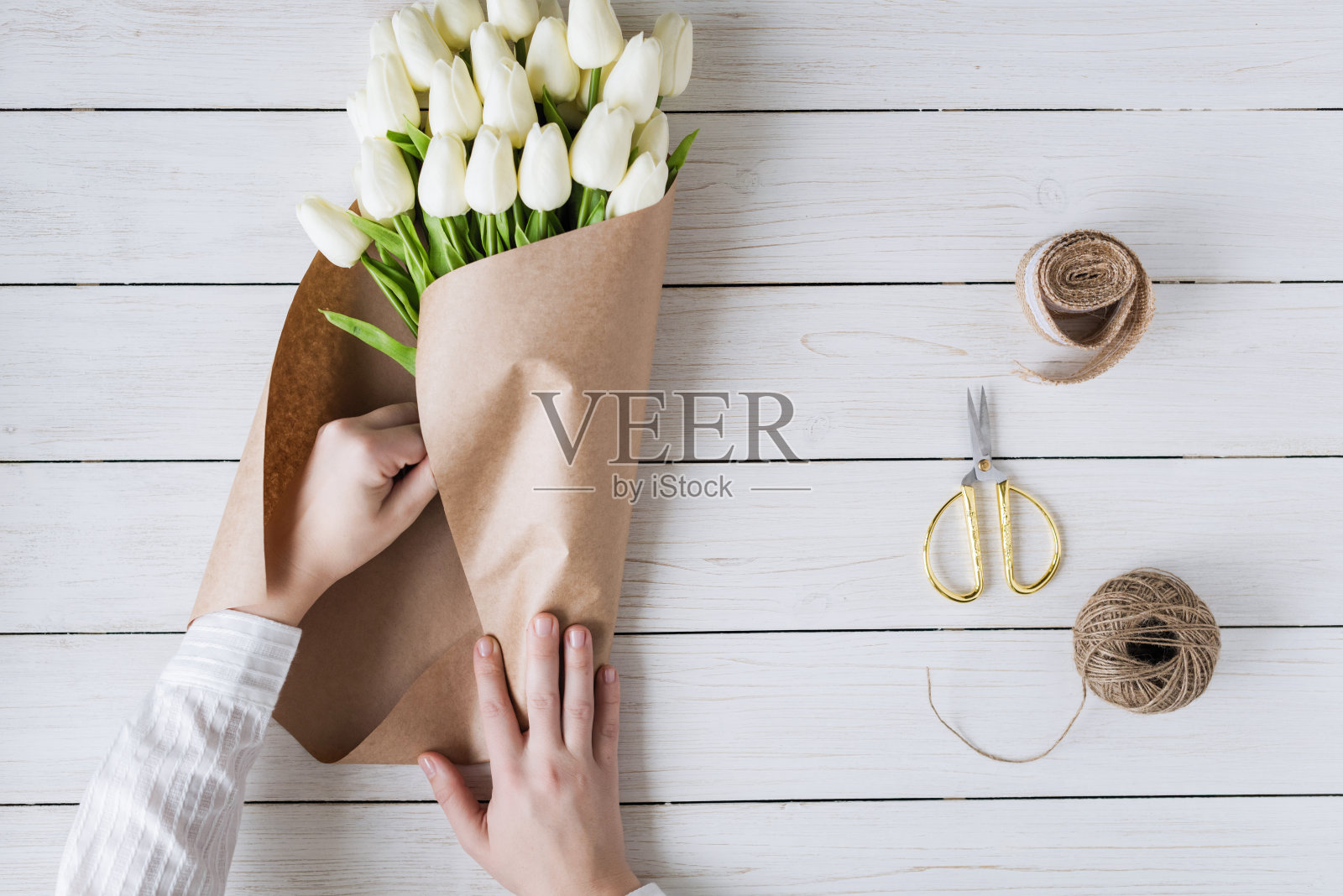 在木桌上，一个卖花的女人正在用包装纸包装一束白色郁金香。平躺照片摄影图片