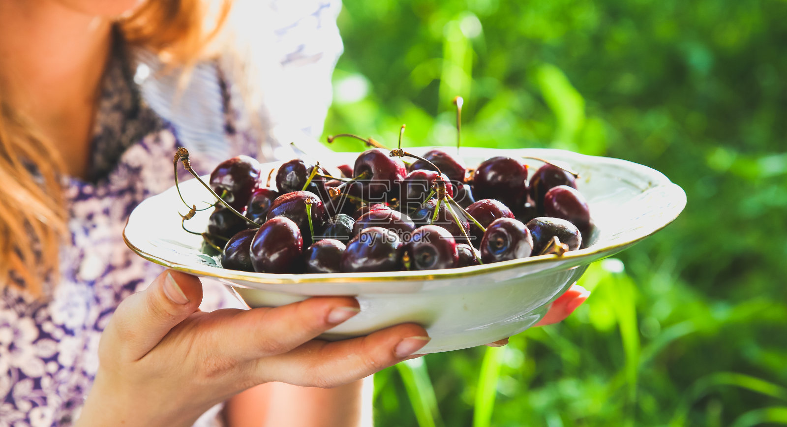 女孩身穿蓝色复古连衣裙坐在草地上，手拿樱桃，乡村夏季水果，健康素食生态食品生活方式概念照片摄影图片