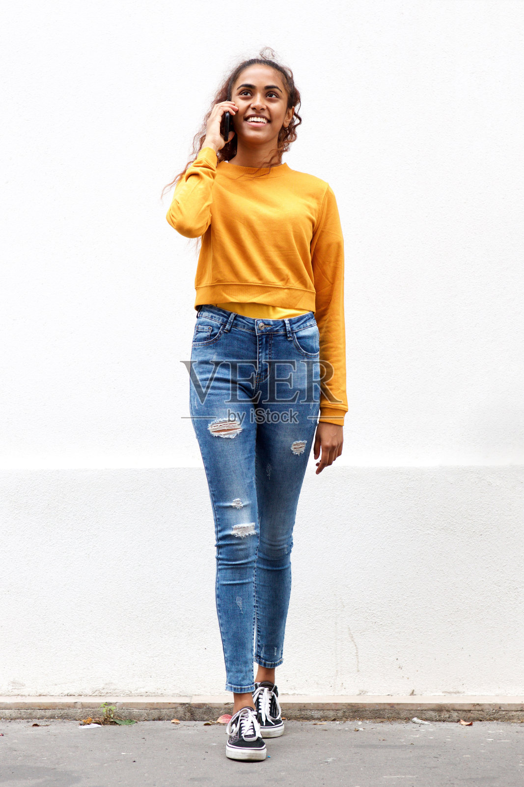 全身美丽的年轻印度女人微笑着白色的墙壁与手机照片摄影图片