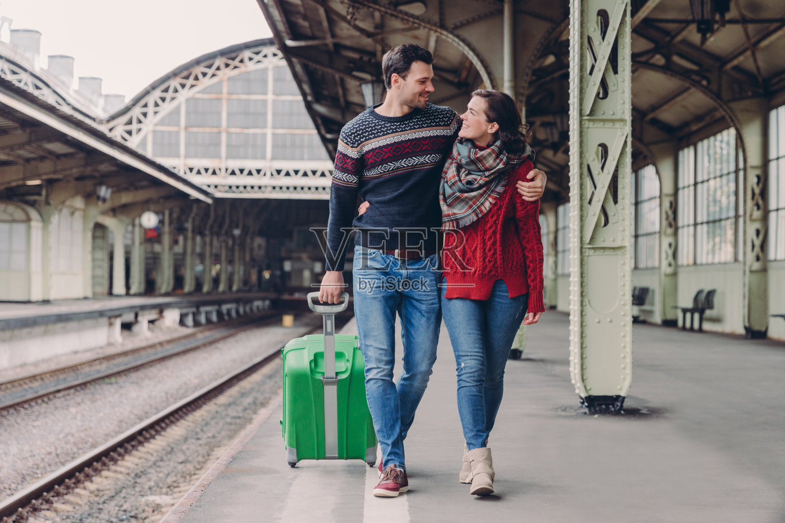 这对年轻的家庭情侣拥抱着，微笑着看着对方，穿着牛仔裤和暖和的毛衣，提着行李箱，在火车站摆姿势，久别重逢。男朋友和女朋友在外面拥抱照片摄影图片
