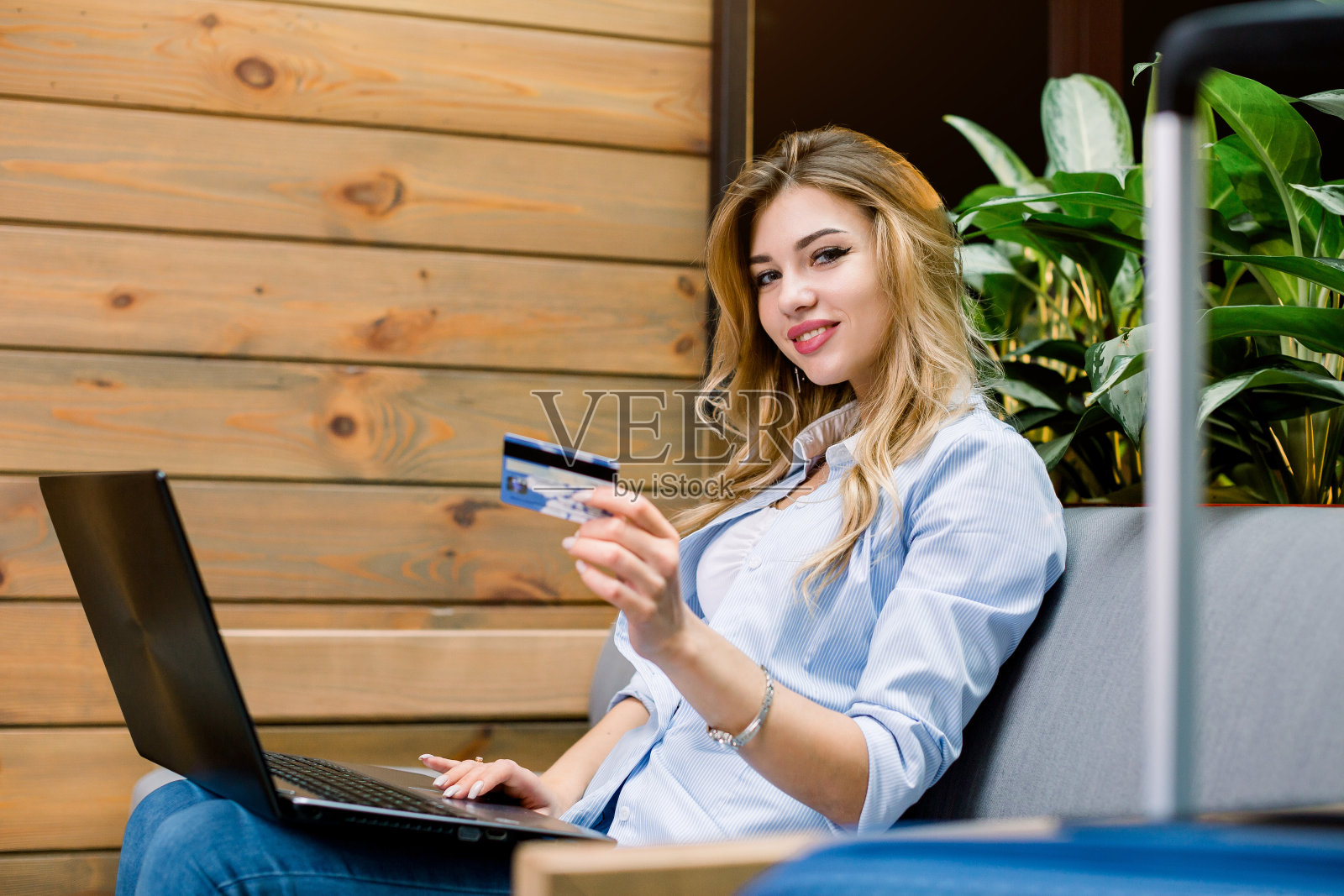 侧面的美丽的年轻女子工作在笔记本电脑和显示信用卡，而坐在现代酒店的沙发上。在线预订、支付、旅游理念照片摄影图片
