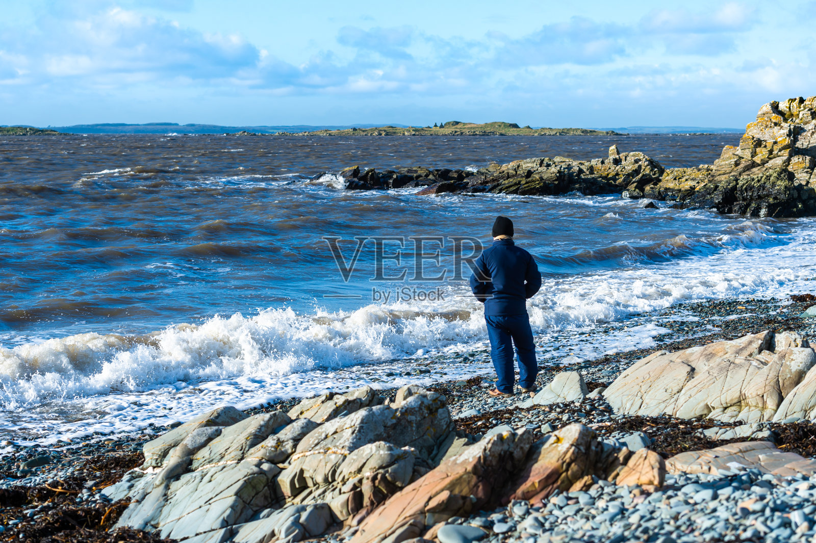 一个老人站在多岩石的海岸上，这是一个晴朗的冬天的早晨，涨潮了。照片摄影图片