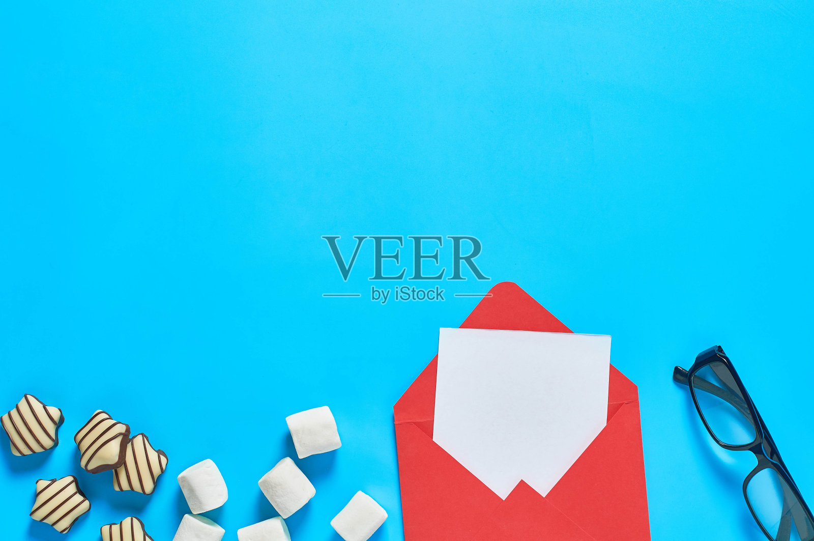 蓝色的桌子上放着散落的糖果、条纹糖果、圆筒状的棉花糖和玻璃边的信封，信封上还有一张白纸。情书的概念。空间的文本。俯视图照片摄影图片