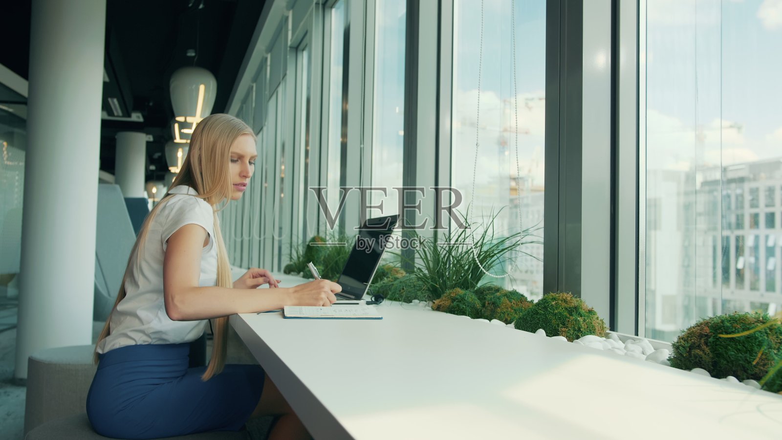 穿正装的女人使用笔记本电脑和文件。女商人坐在现代办公室的窗户旁的桌子旁，一边用笔记本电脑一边在纸上写作照片摄影图片
