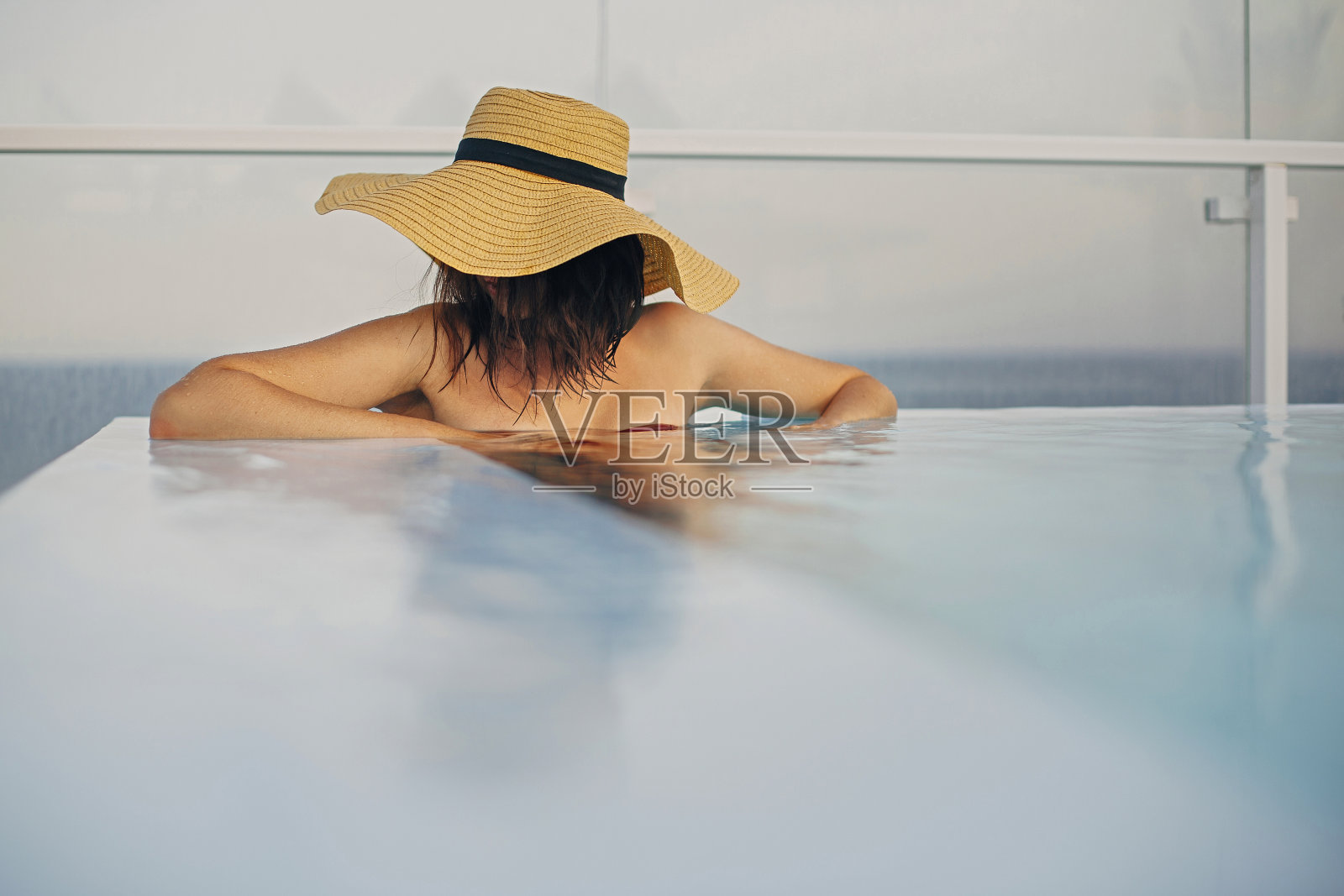 戴着帽子的时尚年轻女子在蓝色泳池中放松身心，享受暑假。夏季热带度假。戴着遮阳帽的女孩在豪华度假胜地度假，在屋顶的游泳池里游泳。留有文字空间照片摄影图片