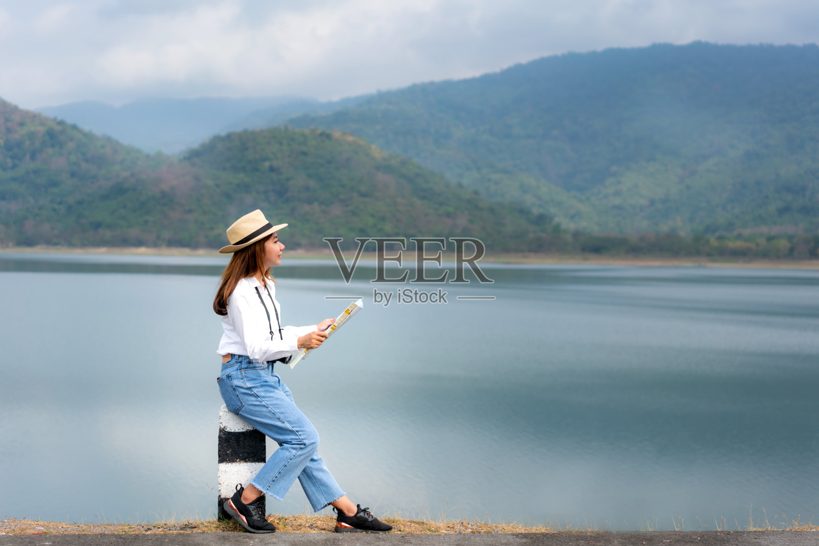 年轻美丽的亚洲女人旅行者与数码相机和地图在手看和高兴看到风景的观点坐在一个湖泊与美丽的山景在泰国。独奏的女人旅行照片摄影图片