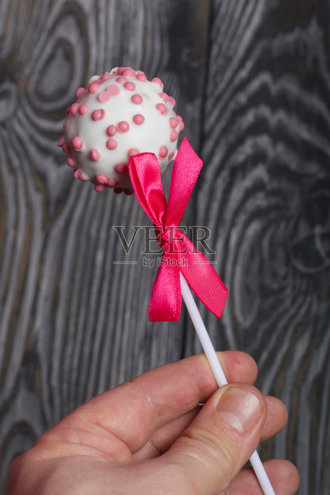 一名男子拿着点缀着粉色糖霜的白巧克力棒棒糖蛋糕。一个粉红色的蝴蝶结系在一根木棍上。以刷过的黑色和白色松木板为背景。照片摄影图片