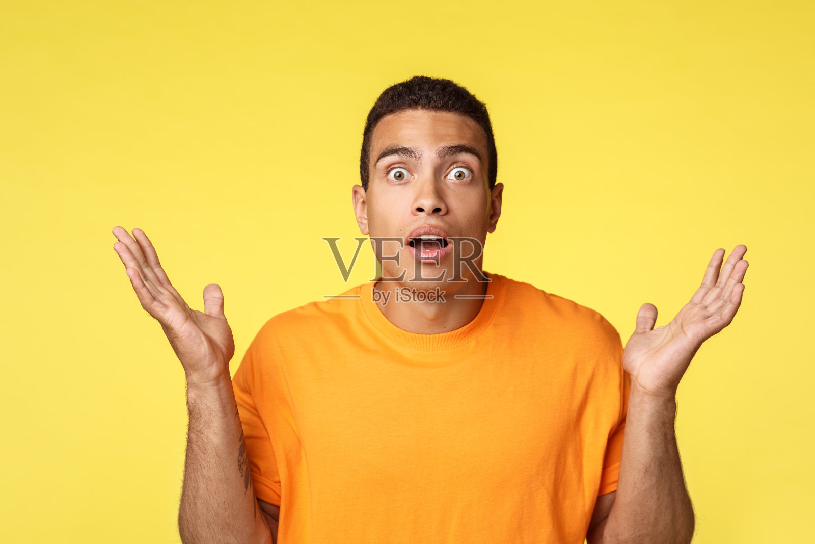 印象深刻和沮丧的年轻男人在比赛中输了赌注，双手向侧面张开，露出沮丧或优柔寡断的手势，张开嘴巴，震惊地凝视着镜头，无语地站在黄色背景前照片摄影图片