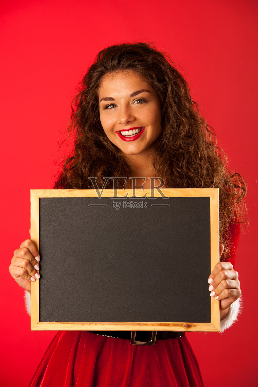 美丽的年轻女子在圣诞老人服装高举黑板在红色的背景照片摄影图片