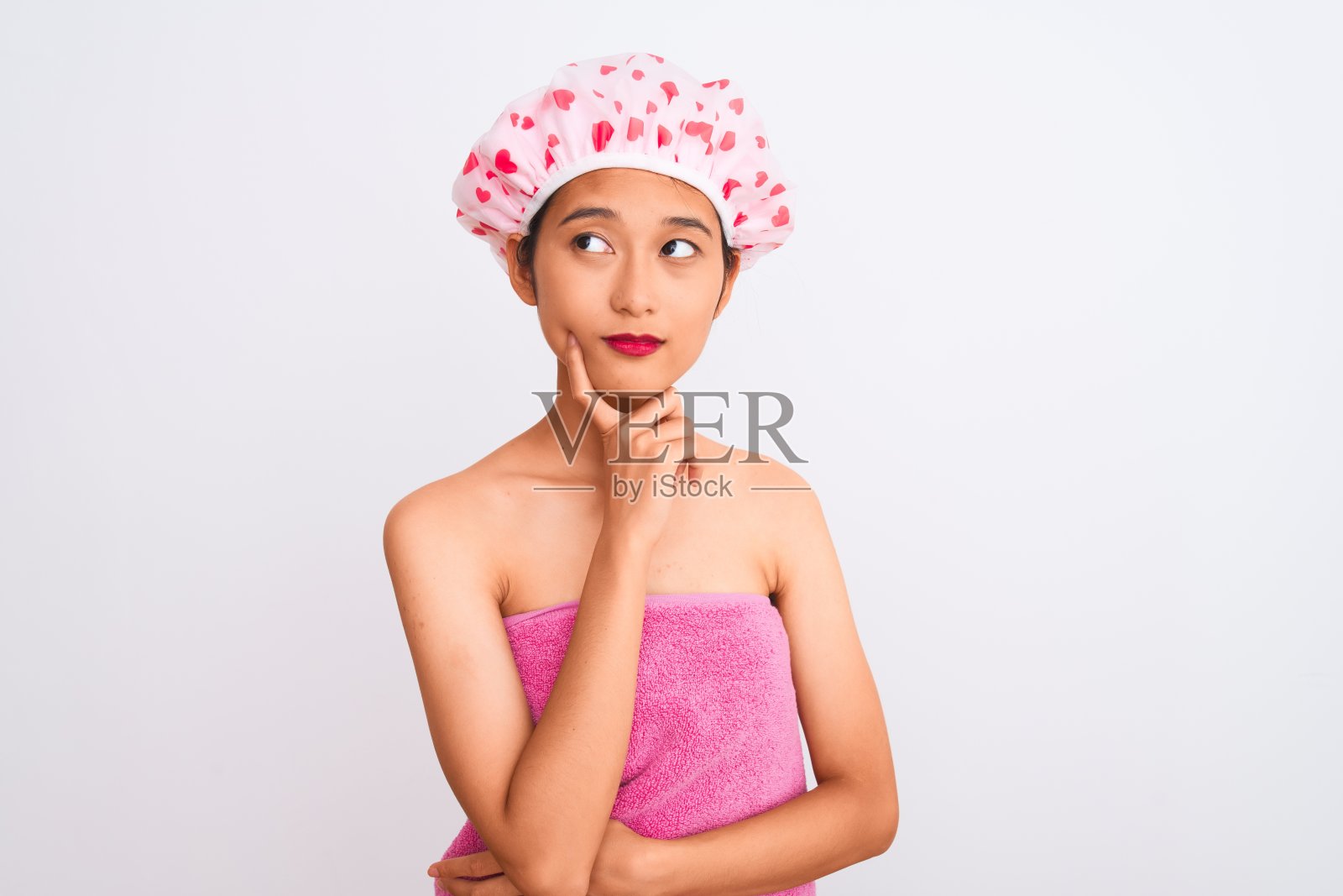 年轻的中国女人穿着浴巾和帽子在孤立的白色背景下，手放在下巴思考问题，沉思的表情。带着沉思的微笑。怀疑的概念。照片摄影图片
