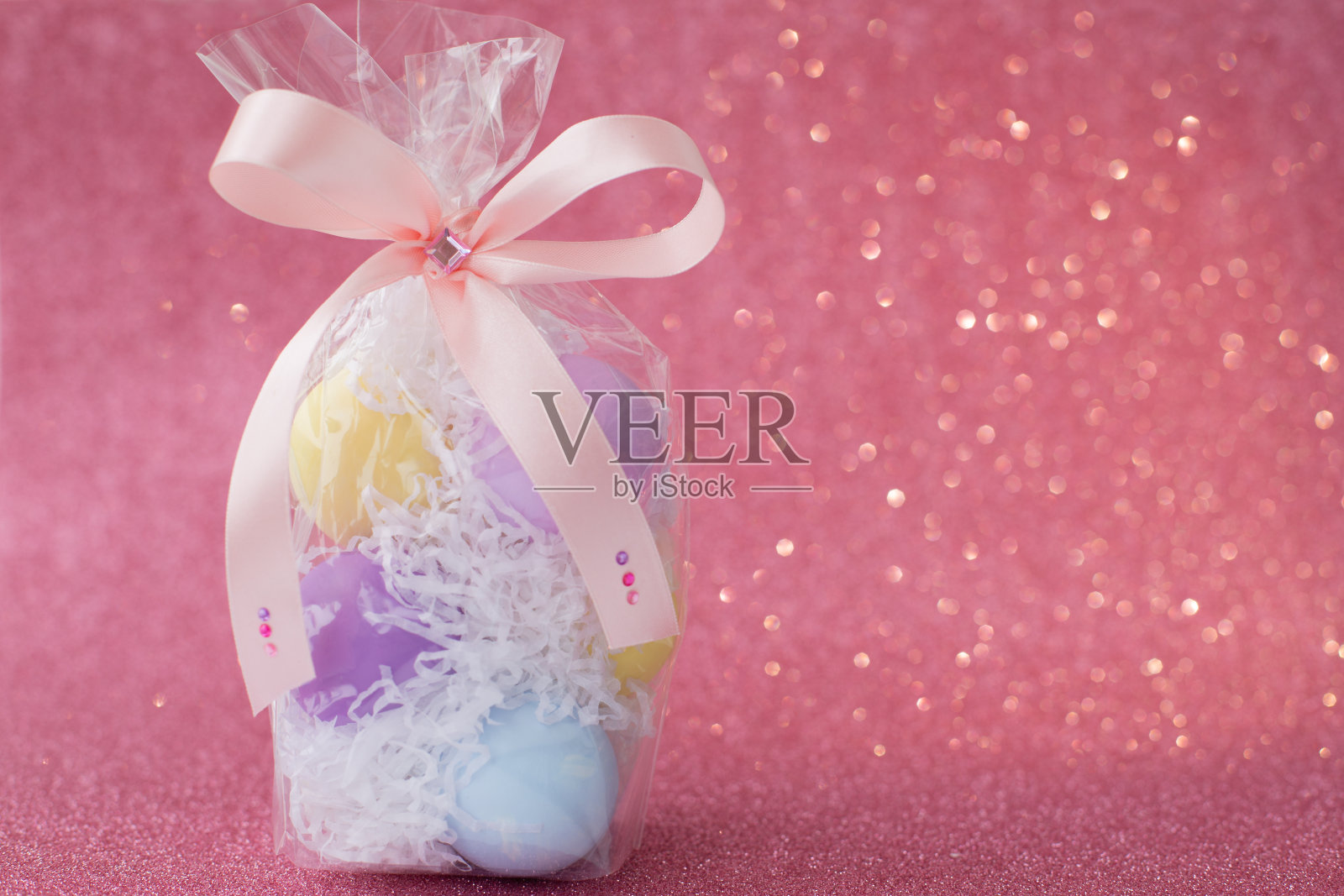 五彩缤纷的紫色、粉色、蓝色、黄色复活节彩蛋，包装在透明的礼品盒中，装饰以蝴蝶结和水钻，以粉红色闪亮的散景为背景。象征庆祝宗教节日照片摄影图片