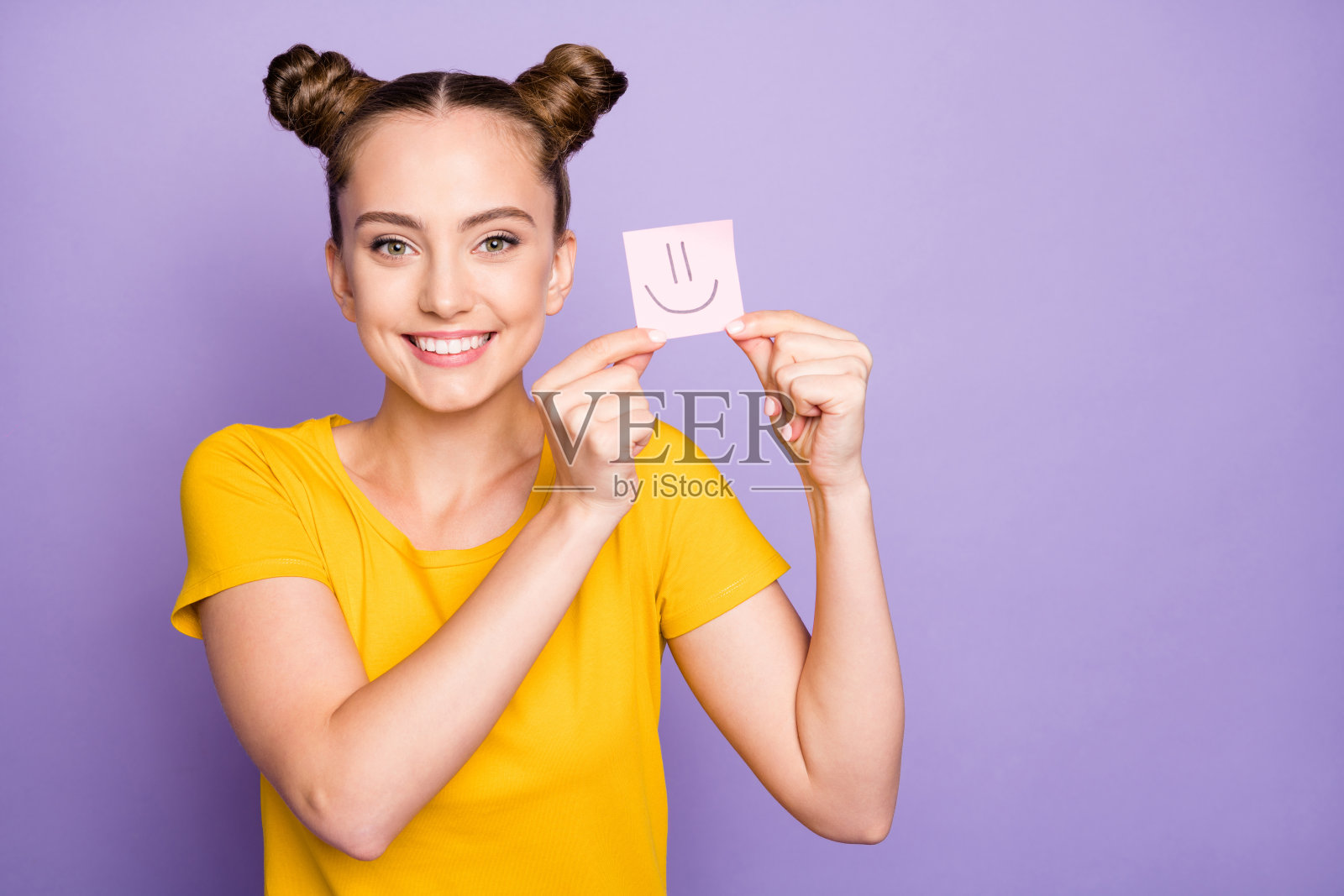 特写：快乐的女士拿着笑脸贴纸，心情愉悦，身穿黄色T恤，背景为柔和的紫色照片摄影图片