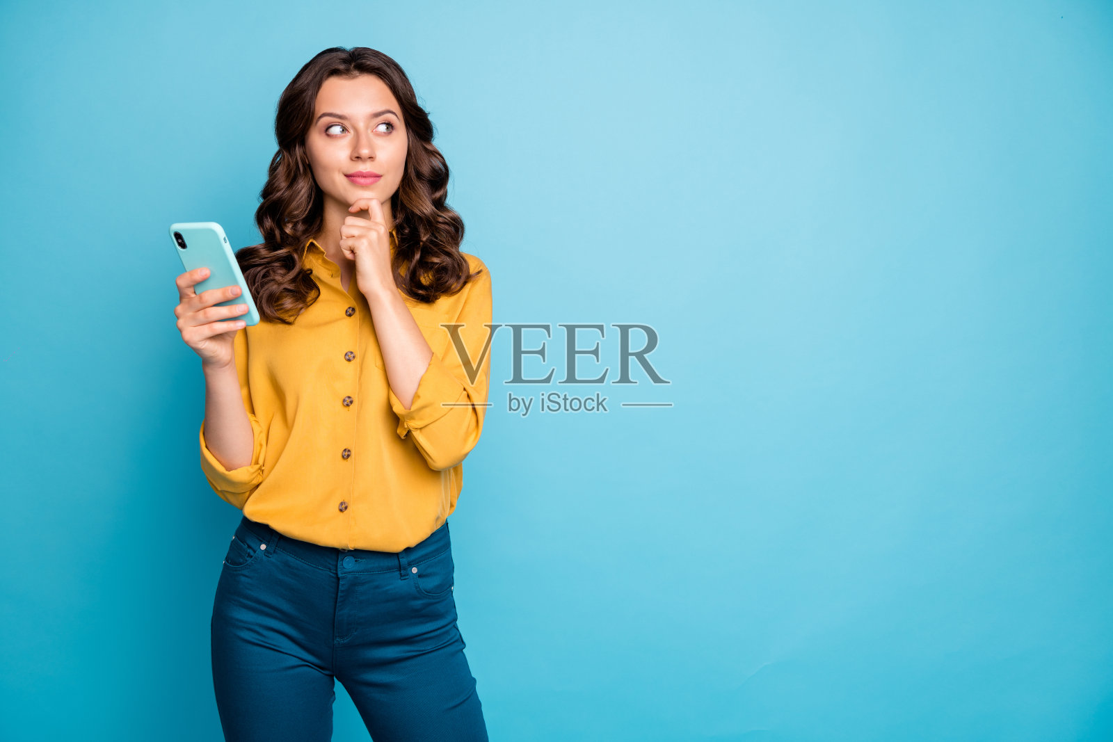 照片漂亮的女士握着电话的手思考创造性的帖子文本的想法看空的空间穿黄色的衬衫裤子孤立的蓝色背景照片摄影图片