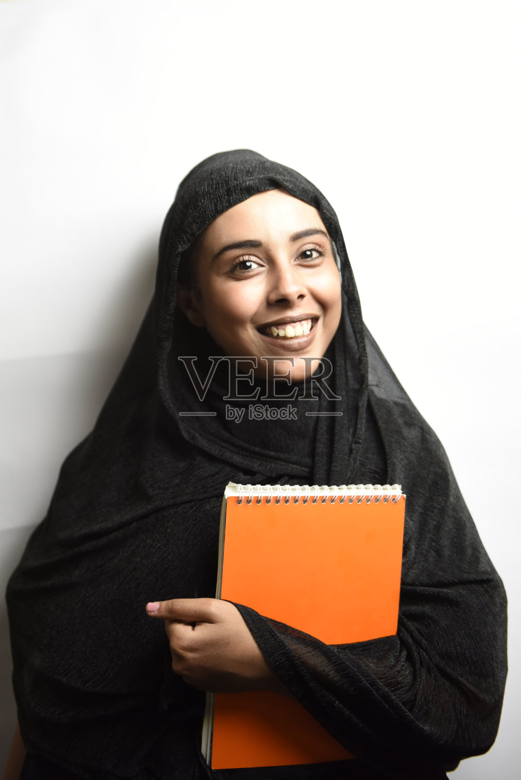 戴着头巾的穆斯林女孩抱着橙色书本，在白色背景上微笑照片摄影图片