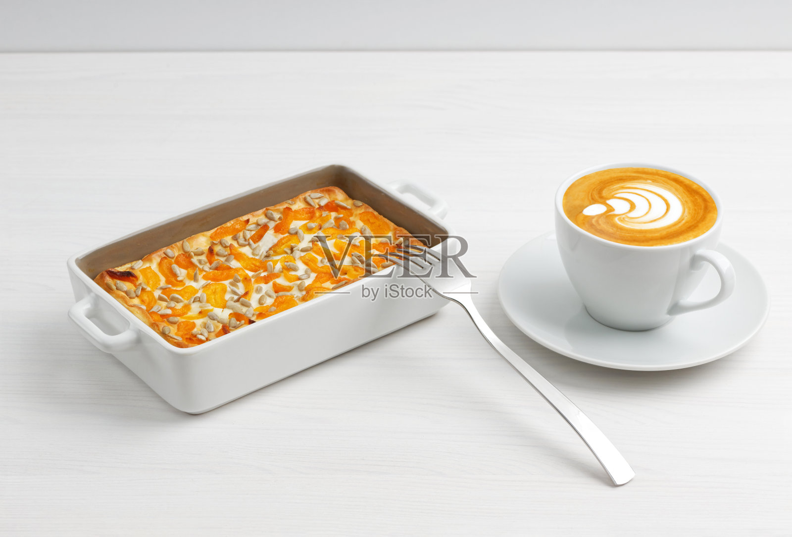 白色木桌上放着一杯咖啡和自制的奶酪砂锅，配上杏干和葵花籽。照片摄影图片