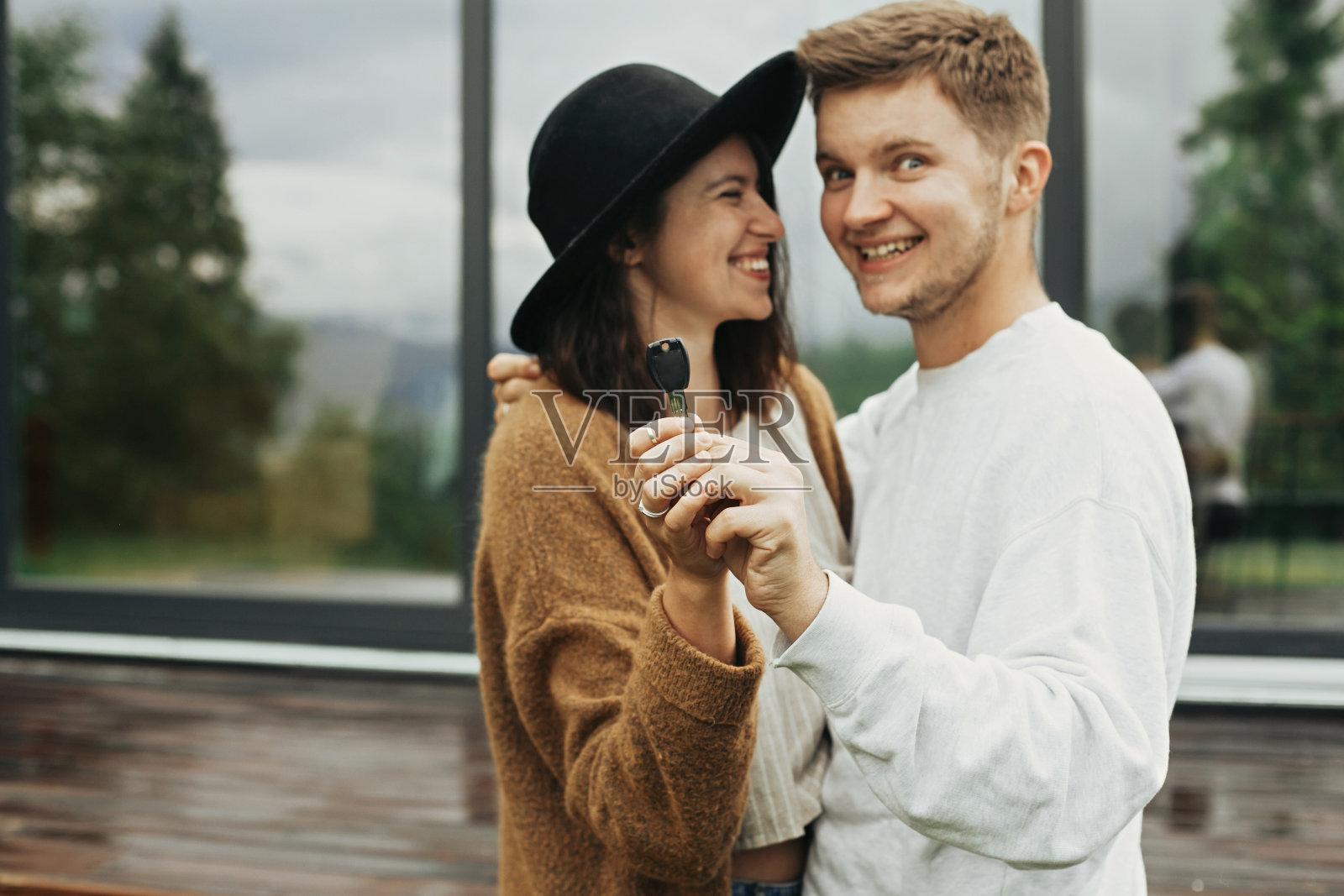 时尚的潮人夫妇在露台和大窗户的背景下，一起拿着他们新家的钥匙。幸福的年轻家庭穿着现代服装庆祝购买第一处房产照片摄影图片