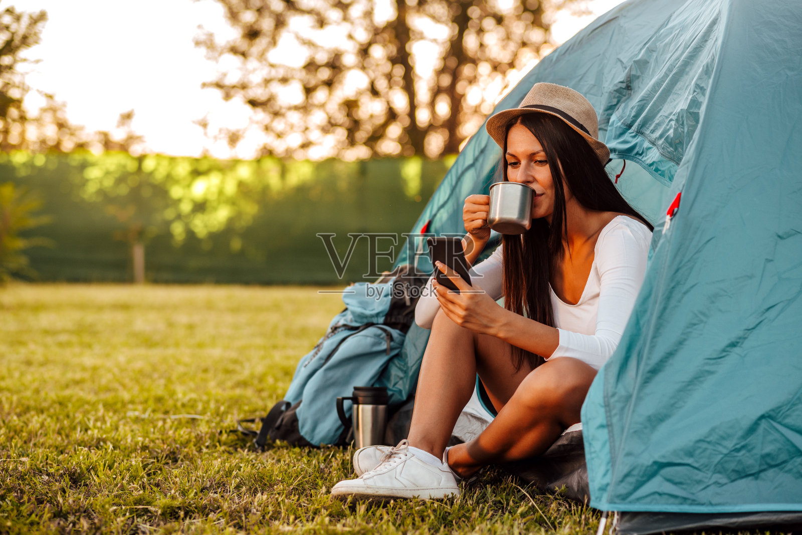 年轻女子在露营时坐在帐篷里喝咖啡看手机的肖像照片摄影图片