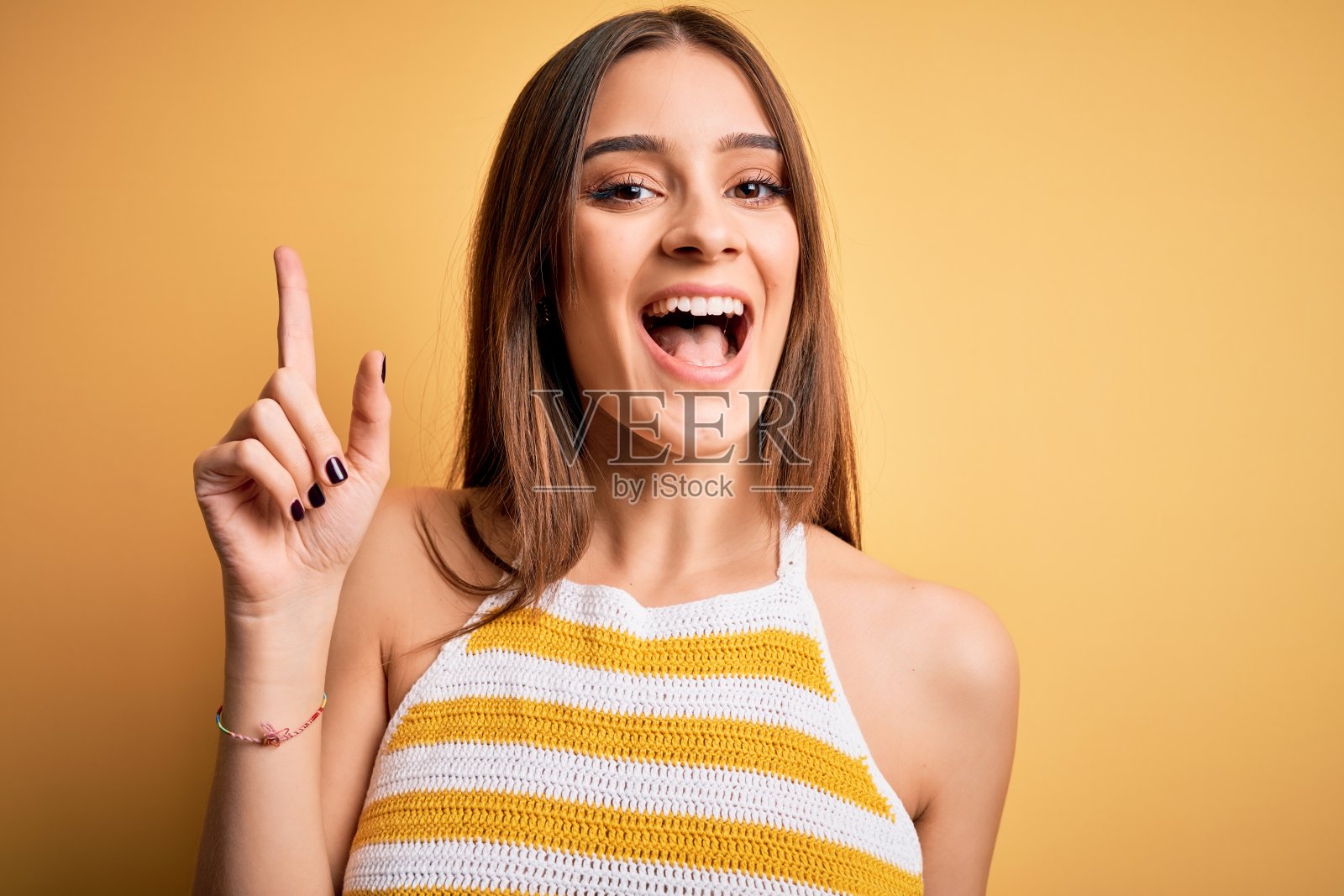 年轻美丽的黑发女人穿着休闲条纹t恤在黄色背景指向手指向上与成功的想法。退出和快乐。第一。照片摄影图片