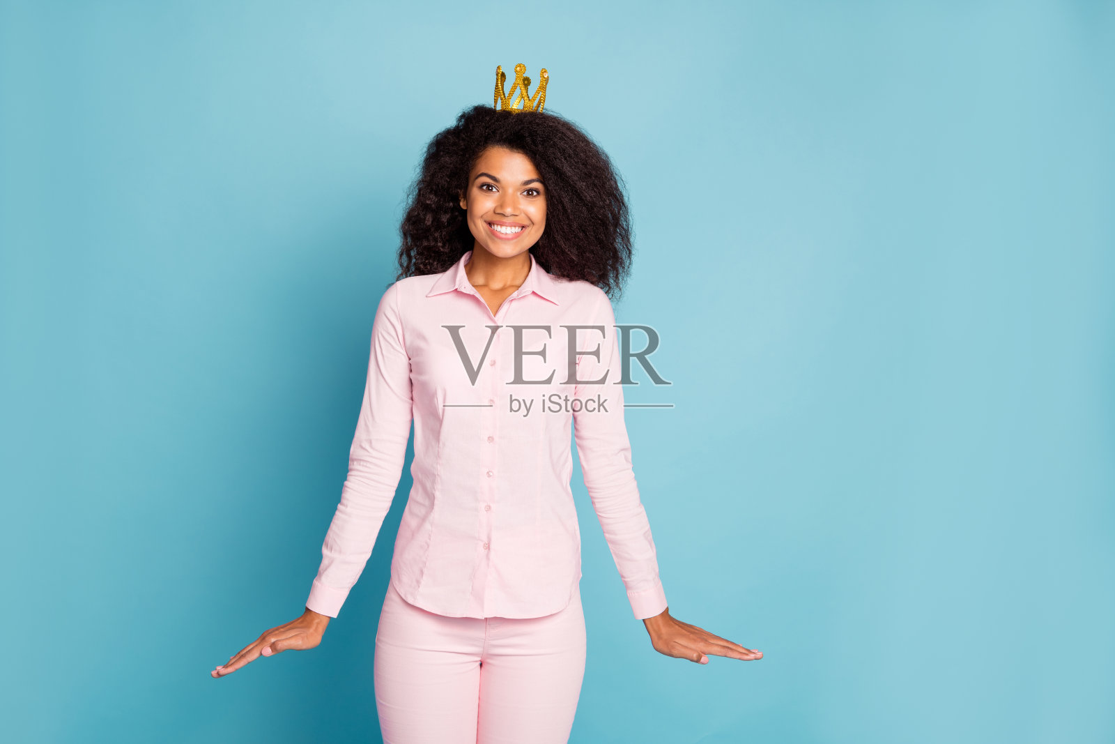 照片惊人的黑皮肤波浪形的女士欣喜地成为舞会皇后害羞的人穿金色皇冠粉红色衬衫裤子孤立的蓝色背景照片摄影图片