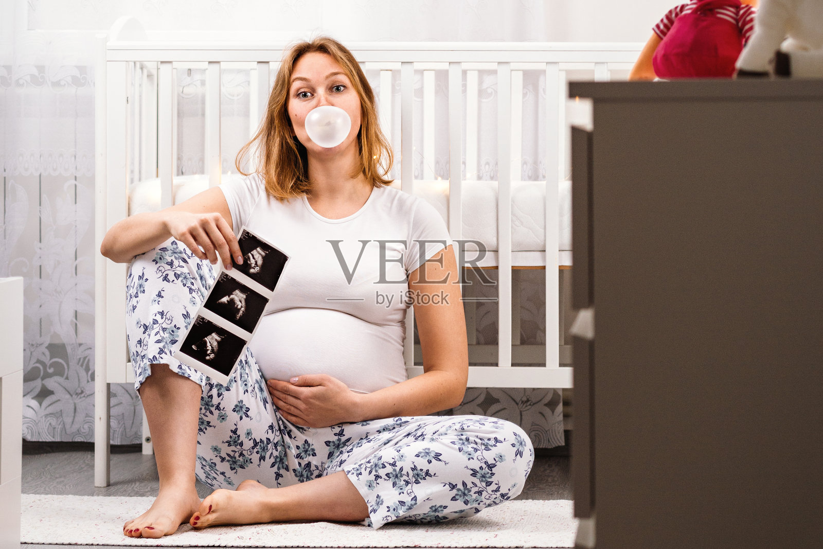 年轻孕妇在地板上拥抱腹部，吹着泡泡糖，拿着超声波图像，母爱概念照片摄影图片