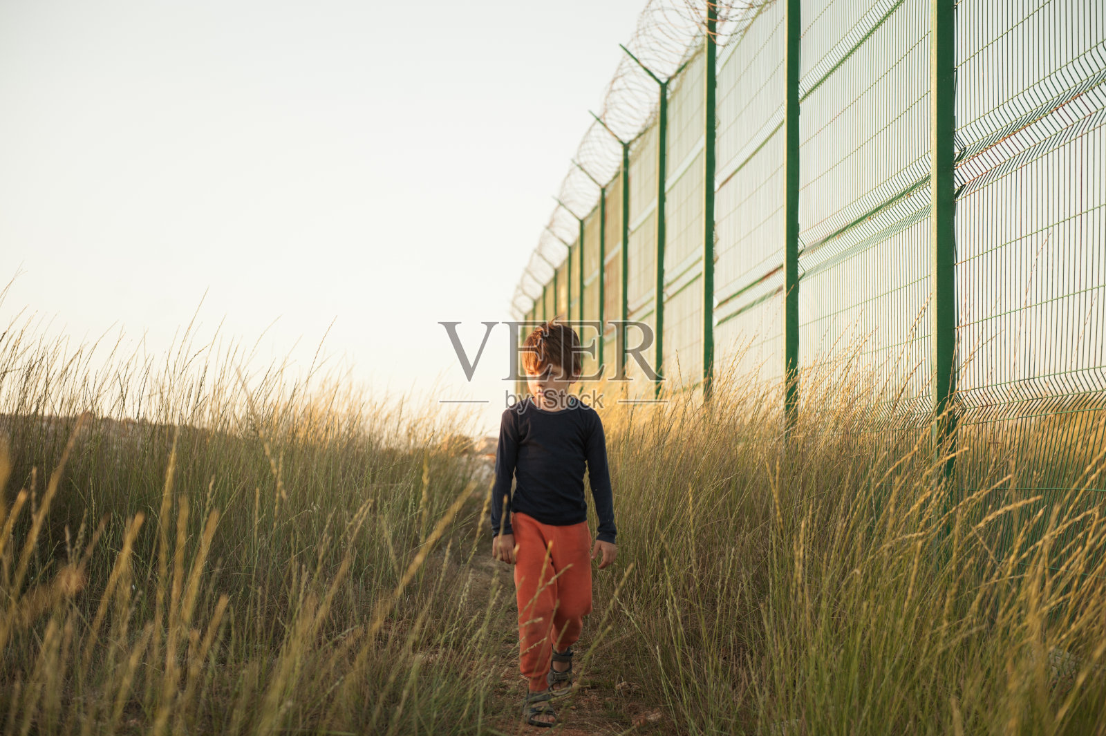 夕阳下，沿着铁丝网围栏行走的可怜白人难民儿童照片摄影图片