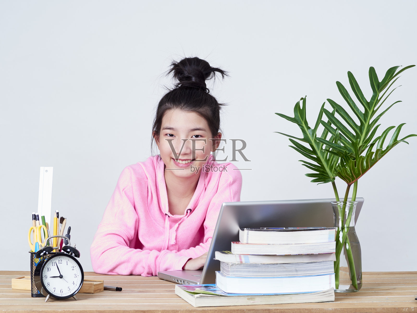 女学生在书桌前使用笔记本电脑，周围堆满了书和闹钟照片摄影图片