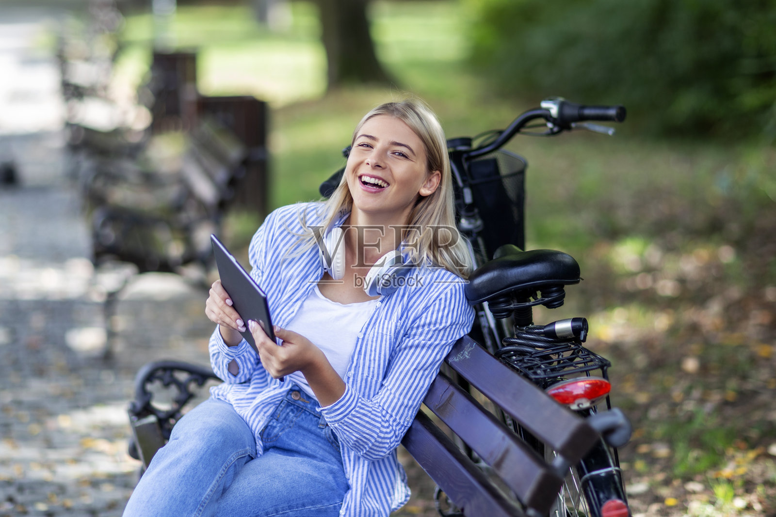 金发女人坐在长椅上，手上拿着平板电脑，脖子上戴着耳机照片摄影图片