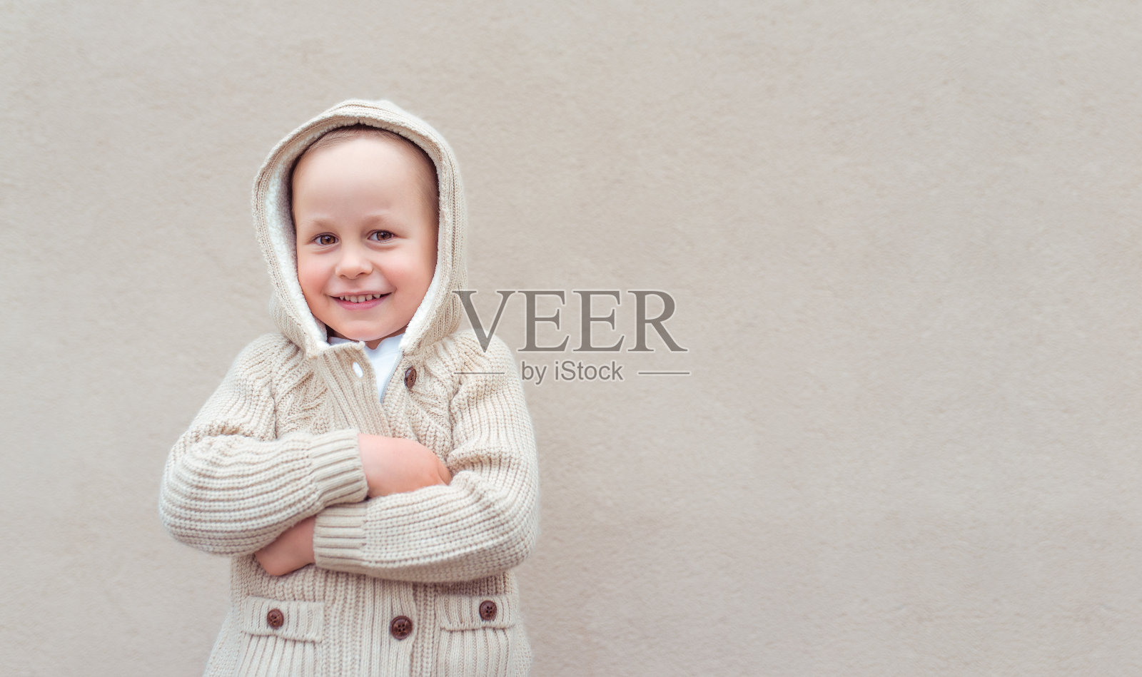小男孩4-5岁，站在米黄色的墙壁上，情绪幸福、快乐、开心、微笑。欧洲婴儿在秋天和春天穿着暖和的衣服。用于复制文本的空闲空间。带兜帽的针织毛衣。照片摄影图片
