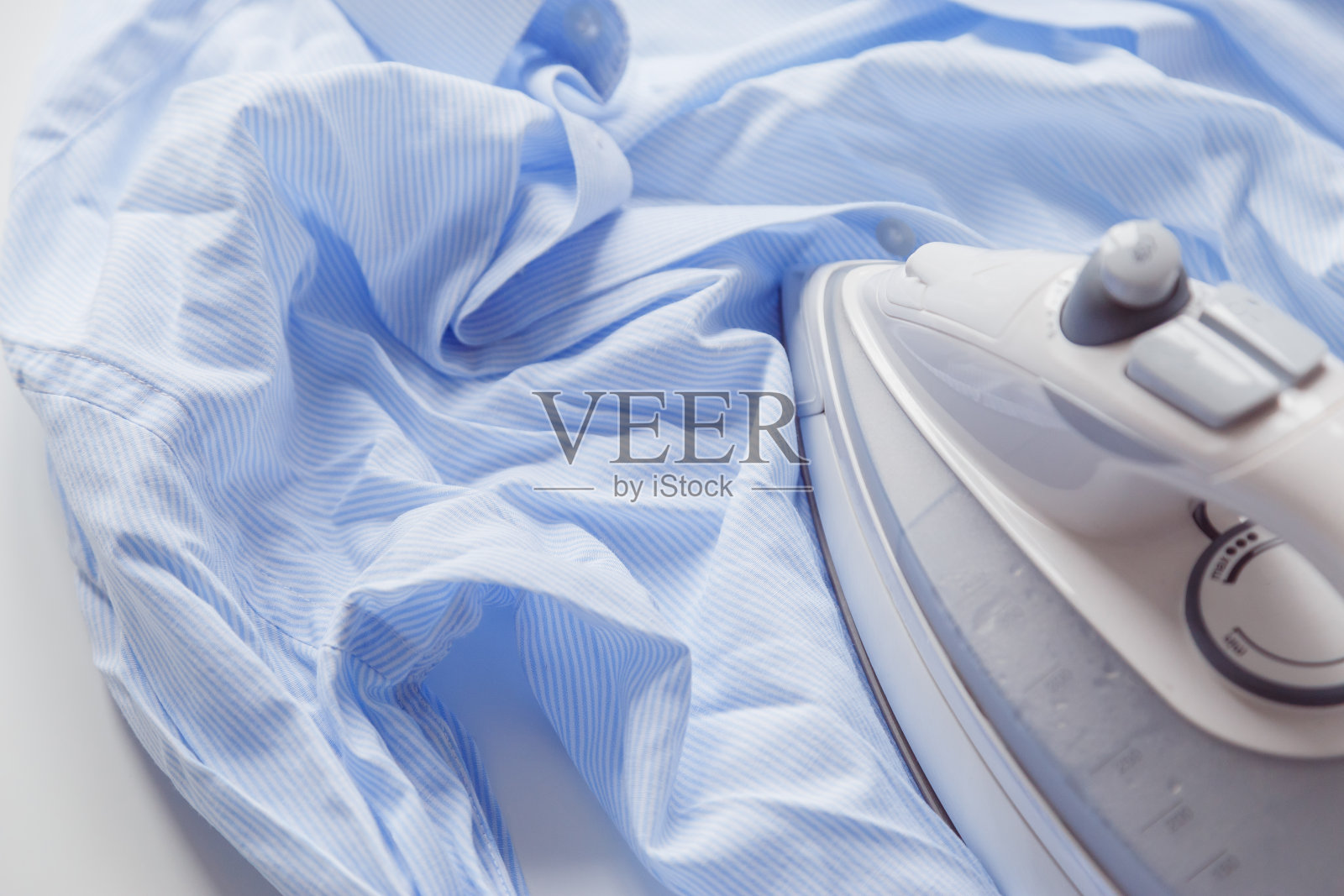 蓝色棉布皱皱衬衫用熨斗熨烫。滚筒烘干机后的洗衣机。照片摄影图片