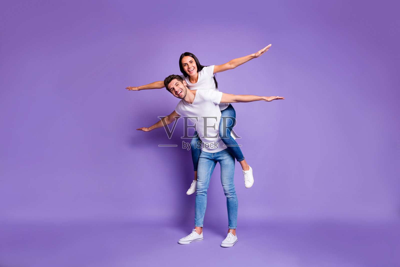 全身尺寸的照片，欢快，兴奋，积极的夫妇与她骑在他的背上，双臂在白色的鞋子t恤牛仔裤牛仔孤立的柔和的颜色紫罗兰背景照片摄影图片