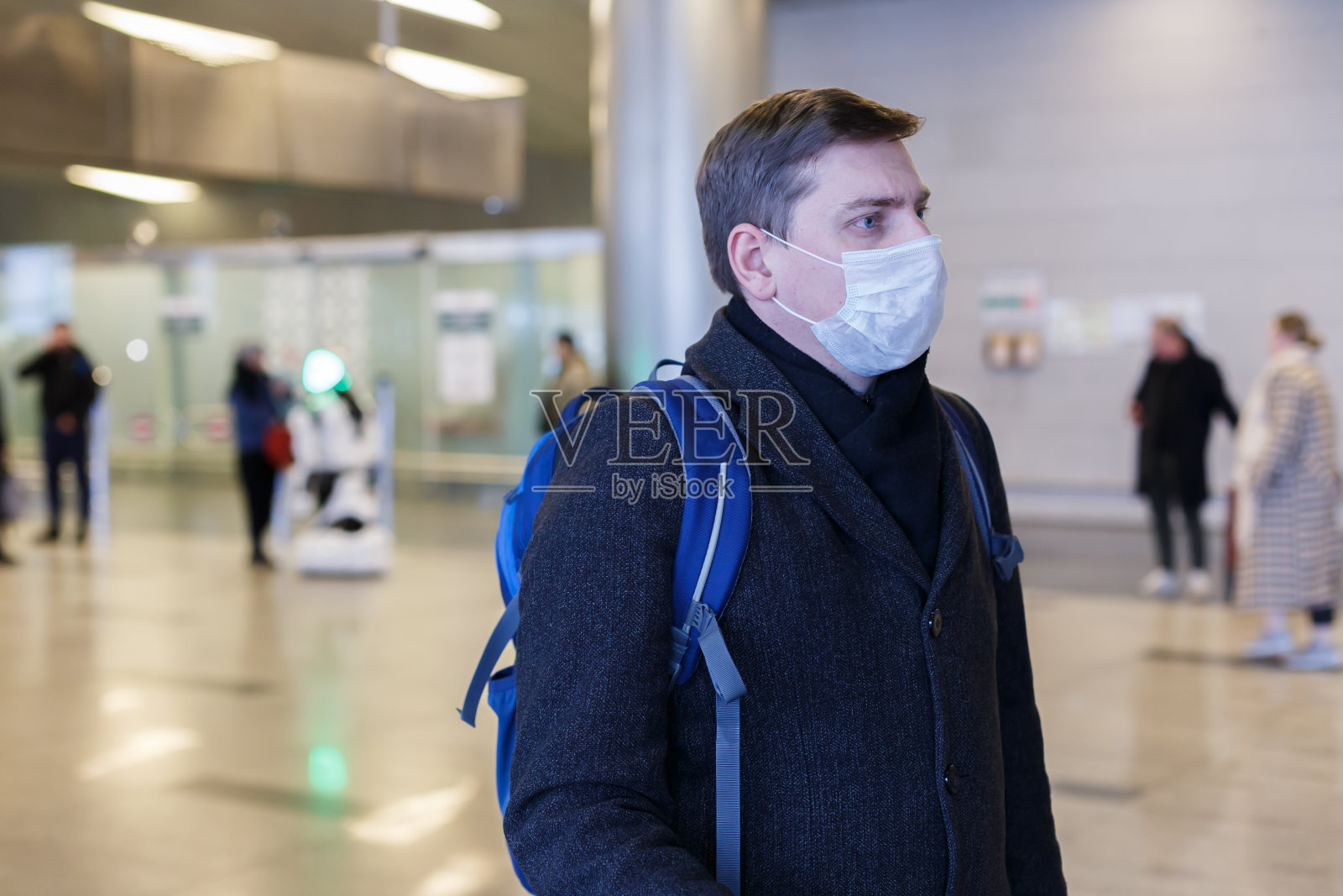 一名身穿灰色外套的年轻欧洲男子，在机场戴着一次性医用防护口罩。害怕危险的新型冠状病毒在变异和传播。蓝色的背包，带轮子的手提箱照片摄影图片
