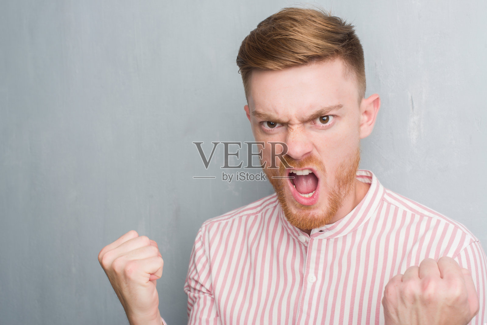 年轻的红发男人越过灰色垃圾墙生气和沮丧的呼喊与愤怒，疯狂的呼喊与举起的手，愤怒的概念照片摄影图片