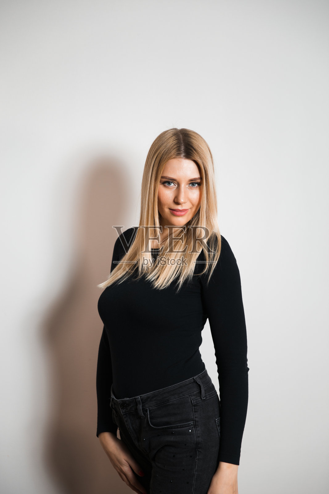 穿着黑色紧身衣和牛仔裤，为相机摄影师摆姿势的迷人长发女子 - 在灰色无缝背景工作室墙上的肖像照片摄影图片