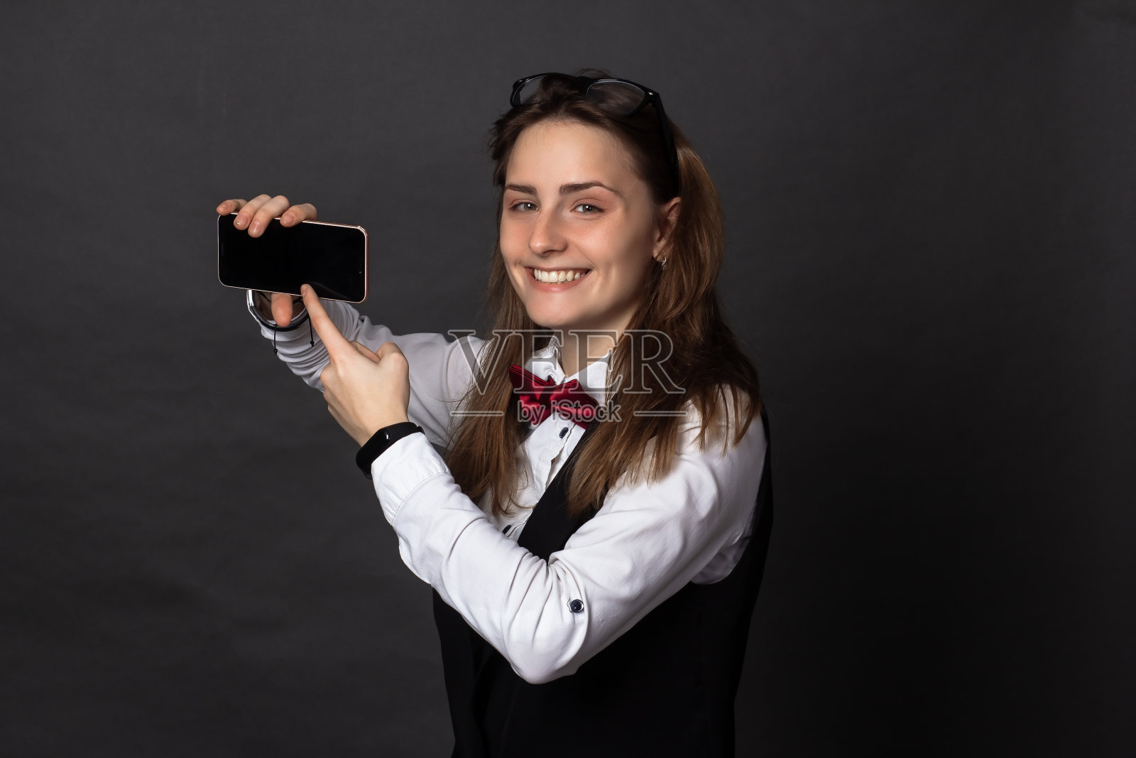 穿着白色衬衫和黑色背心，系着勃艮第领结，戴着眼镜的女孩，快乐地跳跃，用手指指着手机照片摄影图片