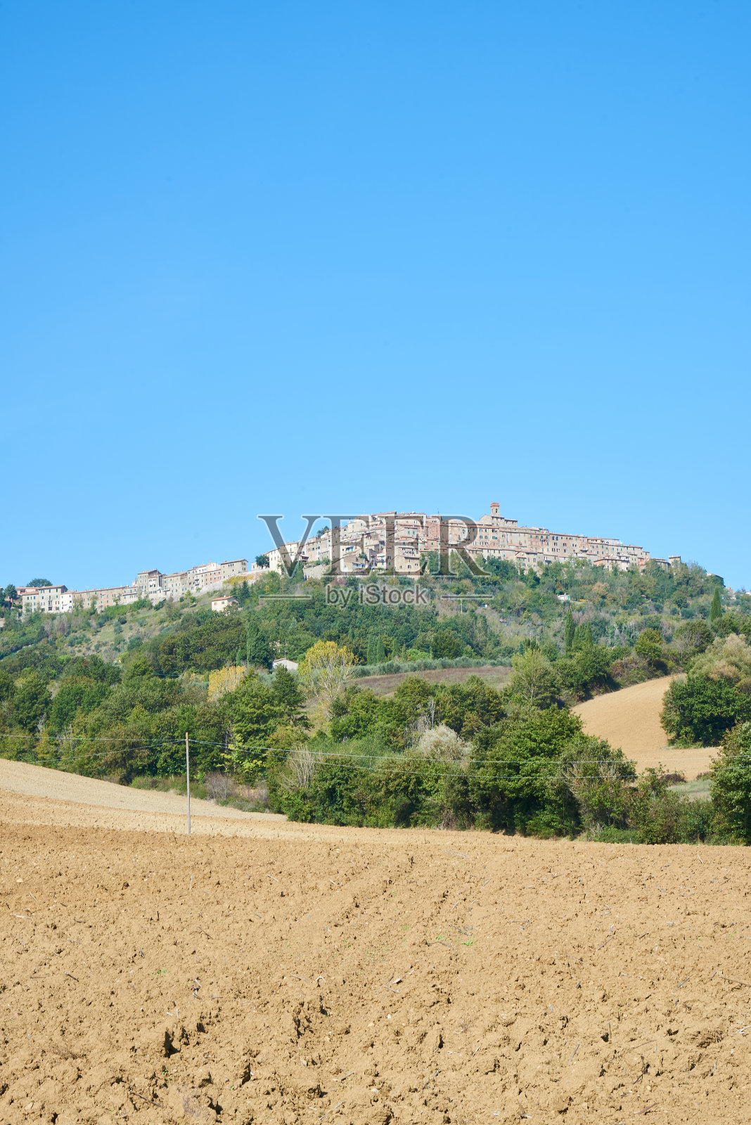 在阳光明媚的日子里，在托斯卡纳Chiusdino附近的乡村，耕地位于绿树和城镇附近的无云蓝天，意大利照片摄影图片