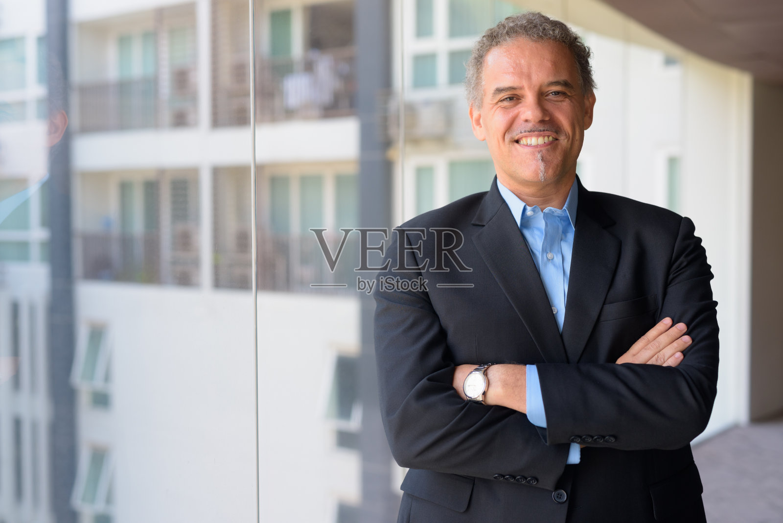 快乐成熟的西班牙裔商人双臂交叉在办公楼的玻璃窗前微笑着照片摄影图片