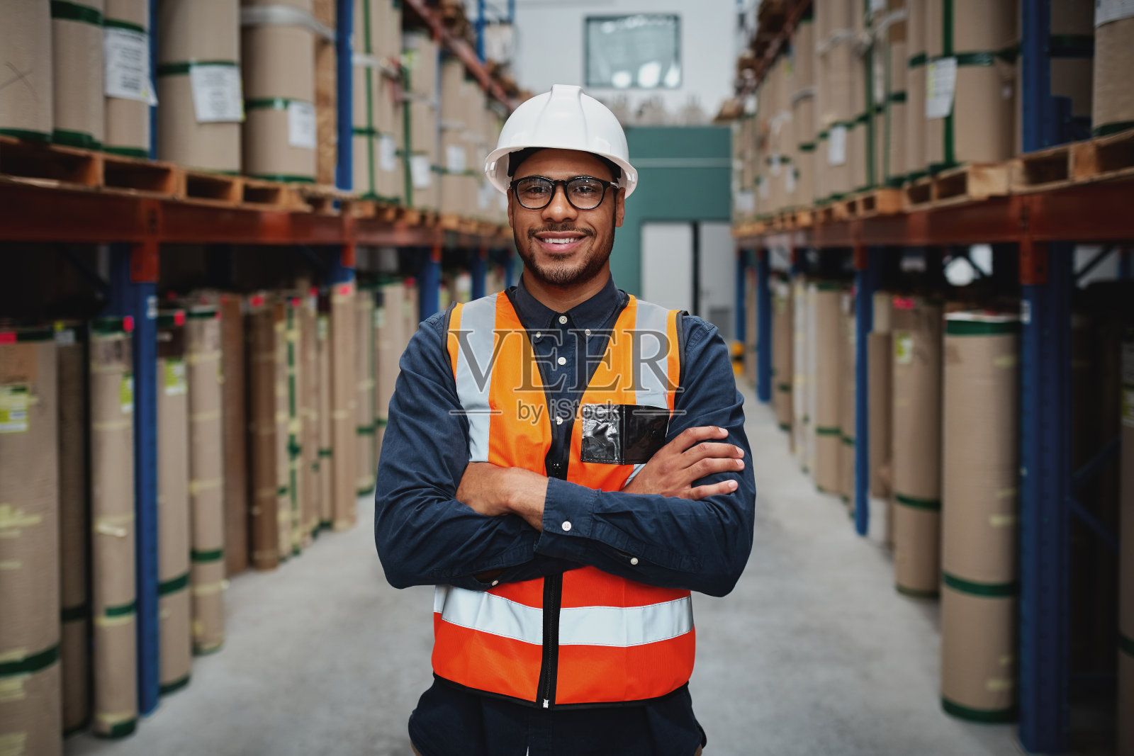 肖像微笑的工人身穿橙色背心和白色头盔，双臂交叉在一个大仓库微笑照片摄影图片