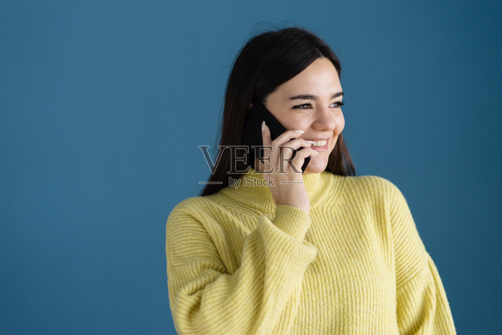 白人女孩的肖像年轻女子摆姿势在蓝色背景墙穿着黄色毛衣用智能手机打电话交谈微笑照片摄影图片