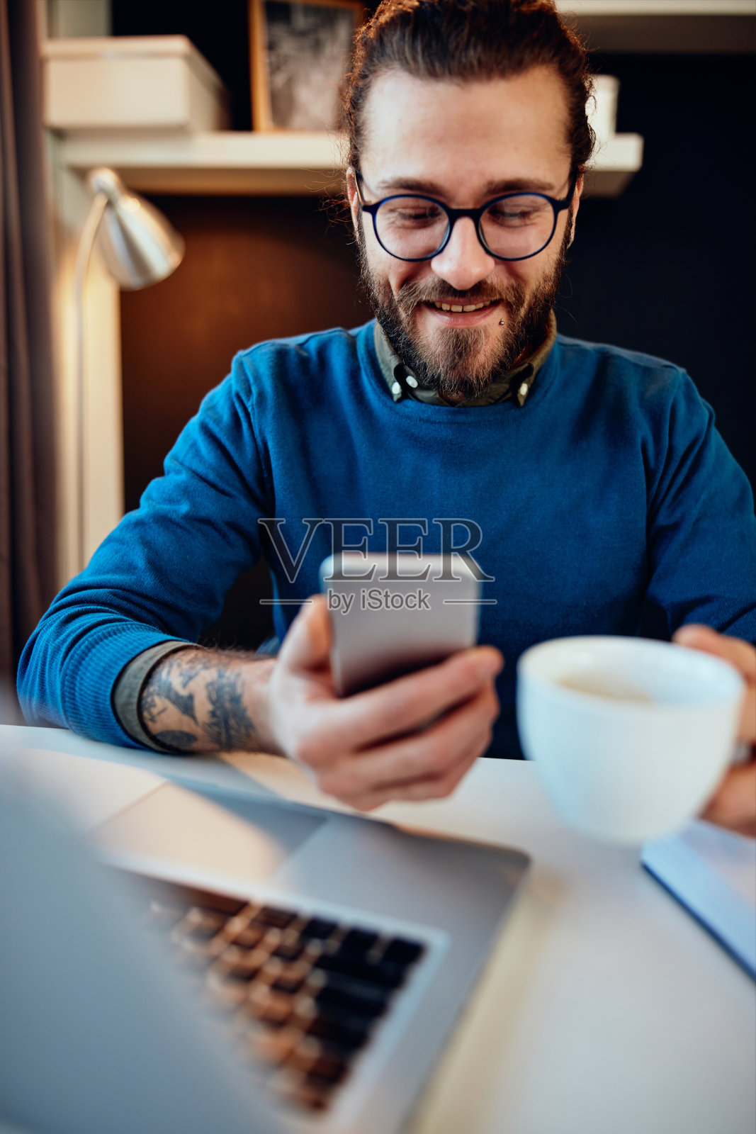 年轻英俊的留着胡子的时髦人士坐在家里的办公室里，拿着咖啡杯使用智能手机照片摄影图片
